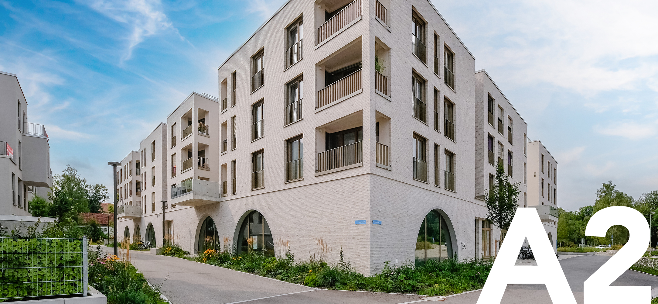 Foto vom Neubau Spötitnger Höfe mit markanten Rundbogenfenstern im Erdgeschoss und grüner Nachbarschaft. In der rechten Bildecke steht A2 als Kennzeichnung des Baufelds