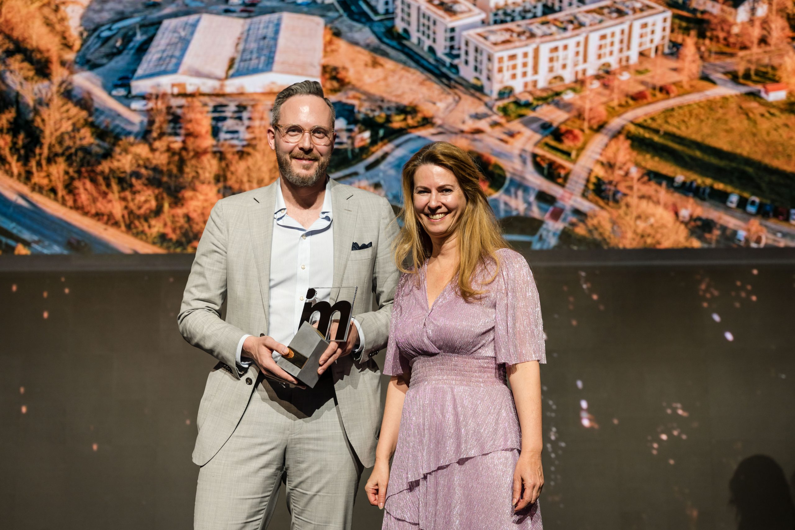 Zwei Personen stehen vor einem Foto eines Stadtquartiers. Eine Person hält einen Preis in der Hand.