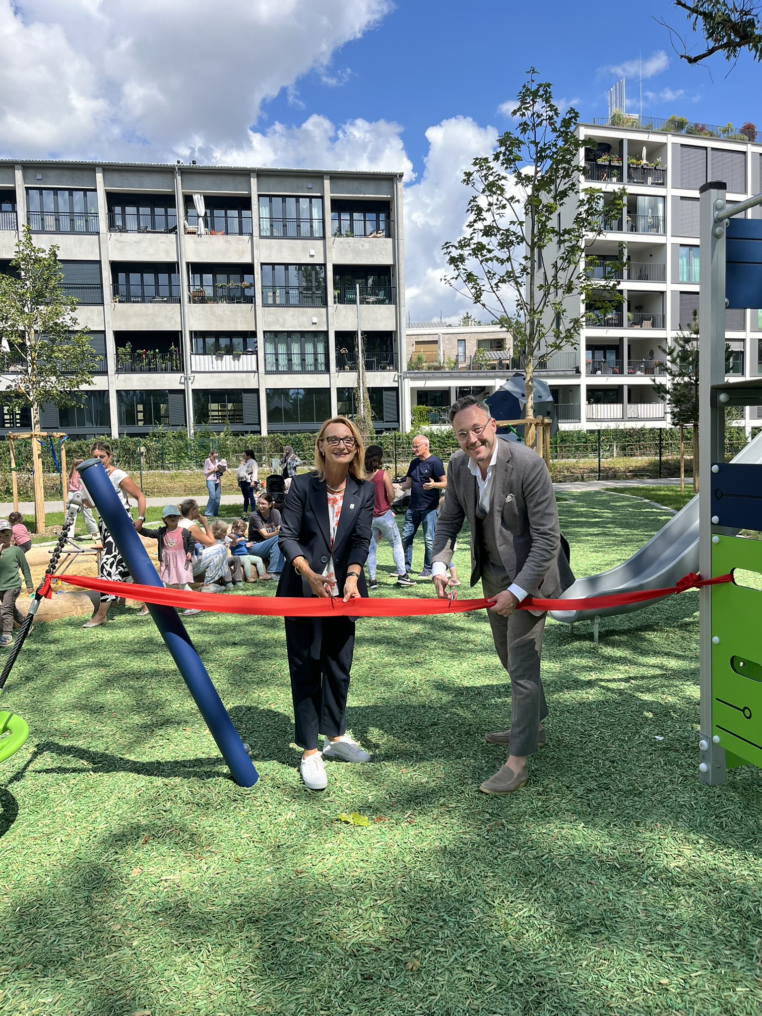 Zwei Personen schneiden ein rotes Band durch. Im Hintergrund sind ein Spielplatz, Häuser und Menschen zu sehen.
