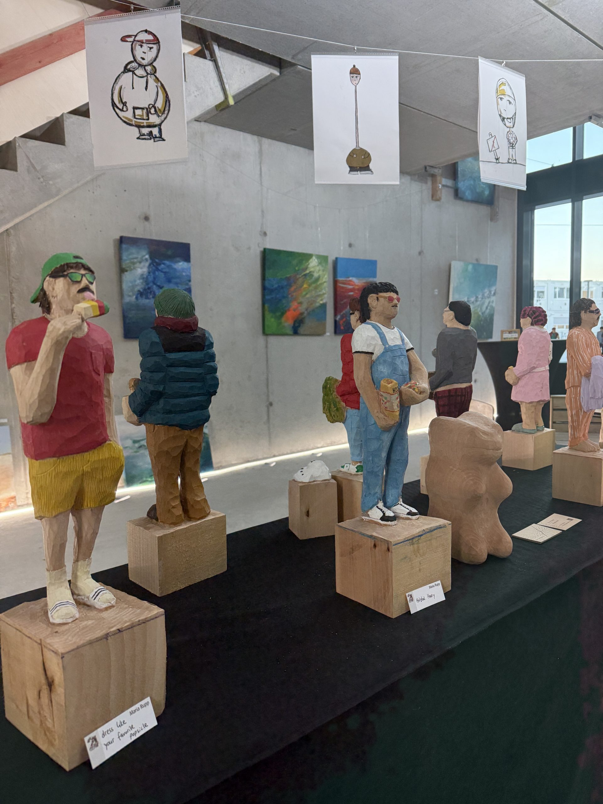 In einem Ausstellungsraum mit Betonwänden sind im Vordergrund bunt bemalte Holzskulpturen zu sehen und im Hintergrund eine Bilderausstellung